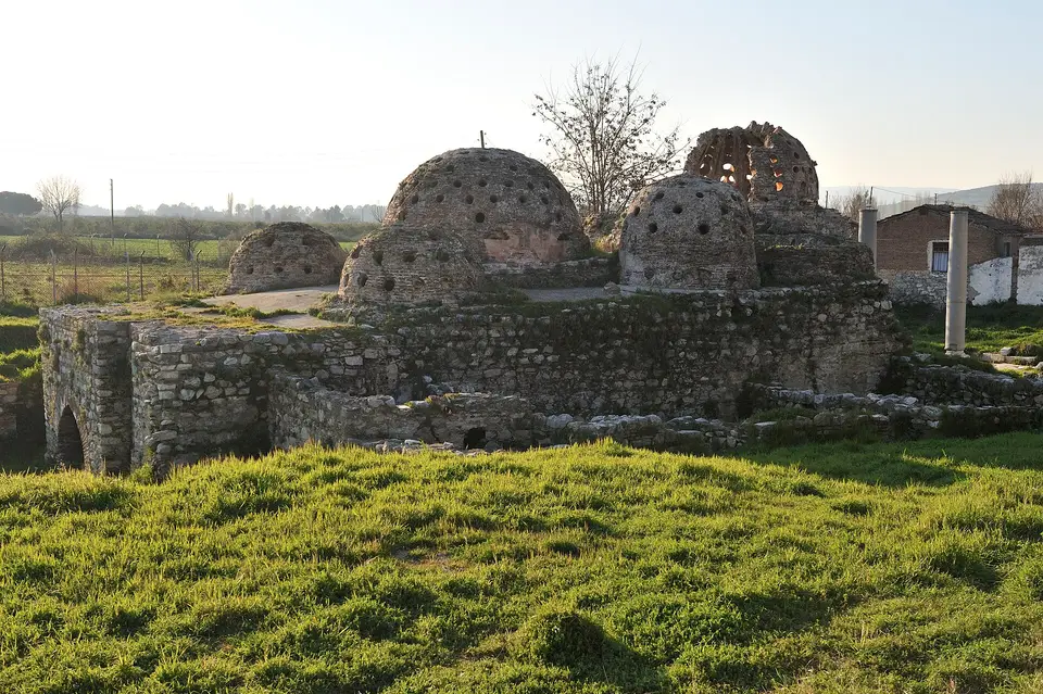 İsa Bey Hamamı’nın yıkık kubbeleri, önde çayır, arkada cami